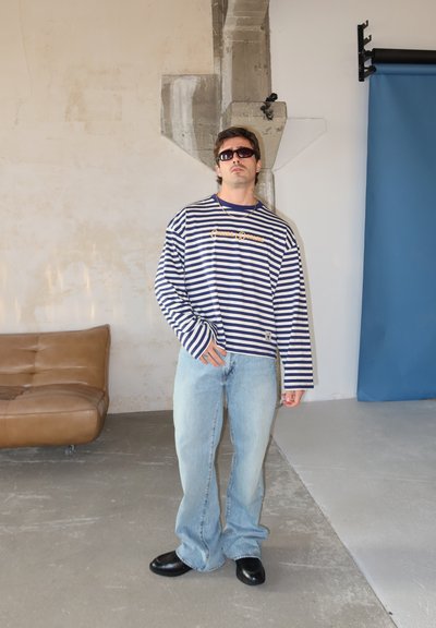 Hombre con camisa de manga larga a rayas, jeans azul claro, gafas de sol y zapatos negros, de pie en una sala industrial moderna con una silla de cuero marrón.