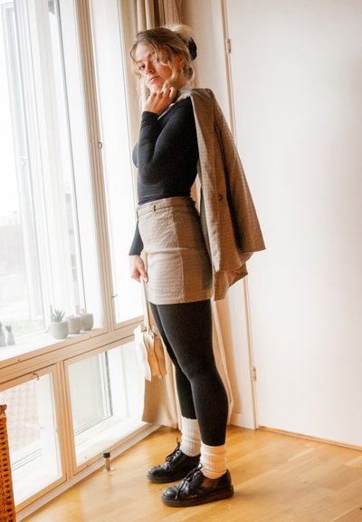 Minifalda de cuadros con cintura alta, top de manga larga negro, medias negras y zapatos de cuero. Blazer marrón claro colgado sobre el hombro. Bolso blanco.