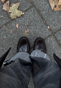 Rjavi usnjeni loaferji z gladkim zaključkom, nošeni z razpokanimi sivimi kavbojkami, na teksturirani tlakovanih tlakovcih posutih z suhim listjem.