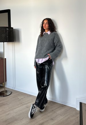 Suéter gris difuso sobre una camisa rosa claro con cuello, combinado con pantalones de charol negro y zapatillas blancas y negras. Fondo minimalista.