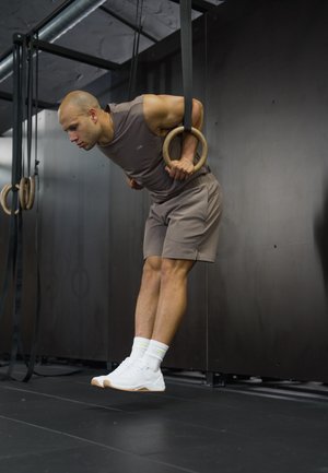 Hombre con ropa deportiva gris realizando un ejercicio de fondos en anillas gimnásticas de madera en un gimnasio interior con paredes y suelo negros.
