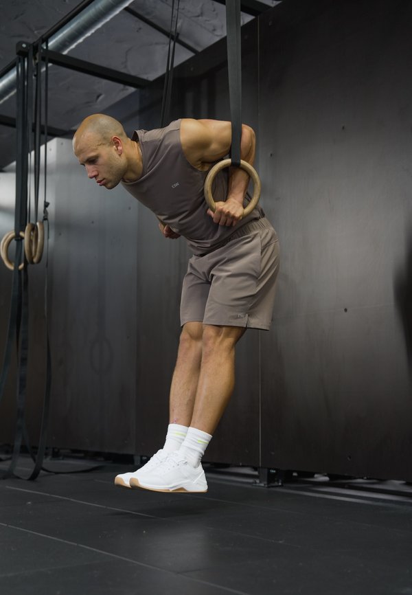 Homme en tenue de sport grise effectuant un exercice de dips sur des anneaux de gymnastique en bois dans une salle de sport intérieure avec des murs et un sol noirs.