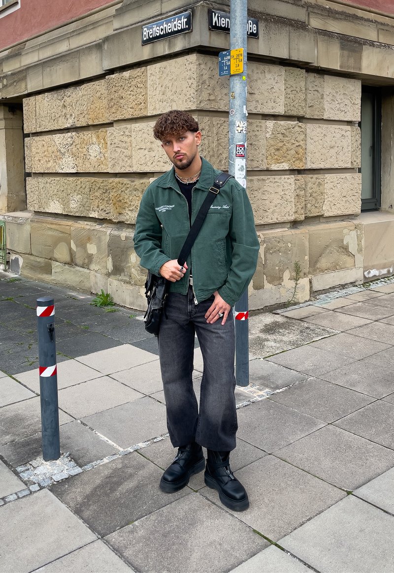 Casaco de lona verde, t-shirt preta, jeans largos pretos, botas plataforma pretas e uma mala de ombro; de pé em pavimento de azulejos com sinais de trânsito.