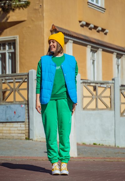 Chaleco acolchado azul brillante sobre un sudadero verde y pantalones de chándal verdes a juego. Un gorro amarillo y unas zapatillas amarillas completan el conjunto.
