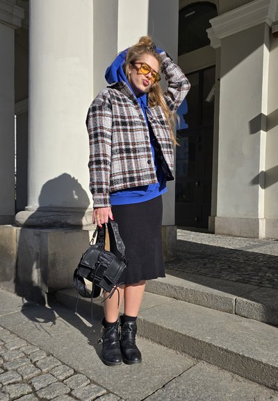 Chaqueta de cuadros con silueta oversized, sudadera azul debajo, falda negra, botines negros y un pequeño bolso negro texturizado.