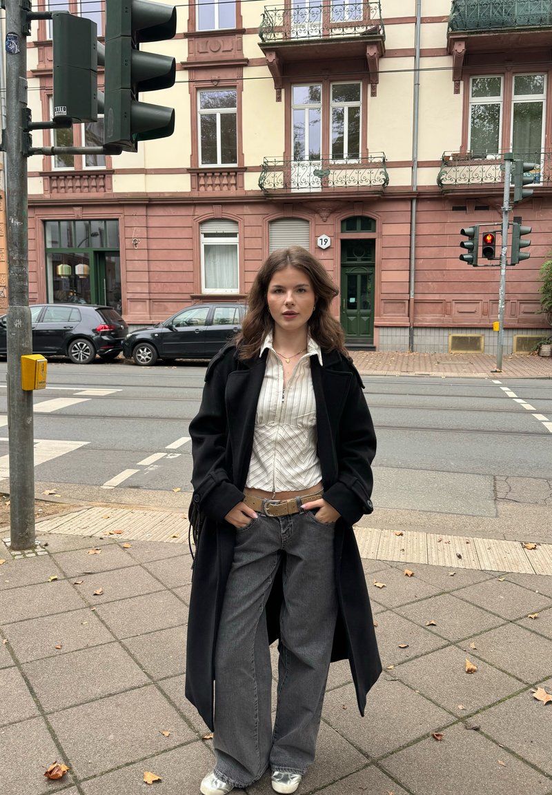 Long manteau noir porté sur une chemise blanche rayée, associé à un jean gris à jambes larges et une ceinture beige. Debout sur un trottoir en ville près des feux de circulation.
