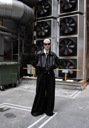 Persona vistiendo una chaqueta bomber de cuero negro sobre una camisa blanca y una corbata negra, con pantalones anchos negros y gafas de sol en un entorno industrial.