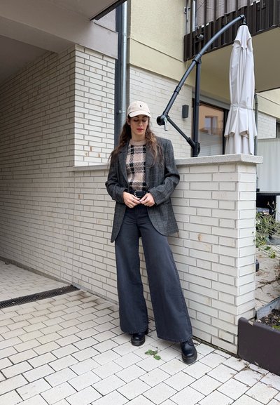 Blazer de cuadros sobre un suéter beige con estampado, combinado con pantalones anchos oscuros y botas negras. Un gorro de color claro completa el conjunto.