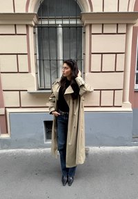 Béžový trenčkot přes černý top, zkombinovaný s tmavě modrými džíny a černými jehlovými lodičkami, stojící proti světle růžové budově.