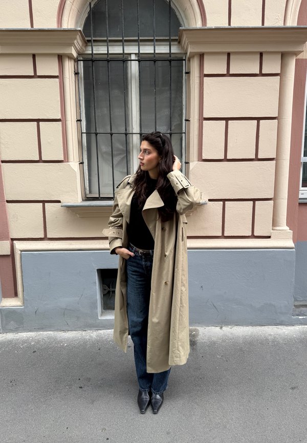 Beige trench coat fekete toppal, sötétkék farmernadrággal és fekete tűsarkú cipővel, egy világos rózsaszínű épület előtt áll.
