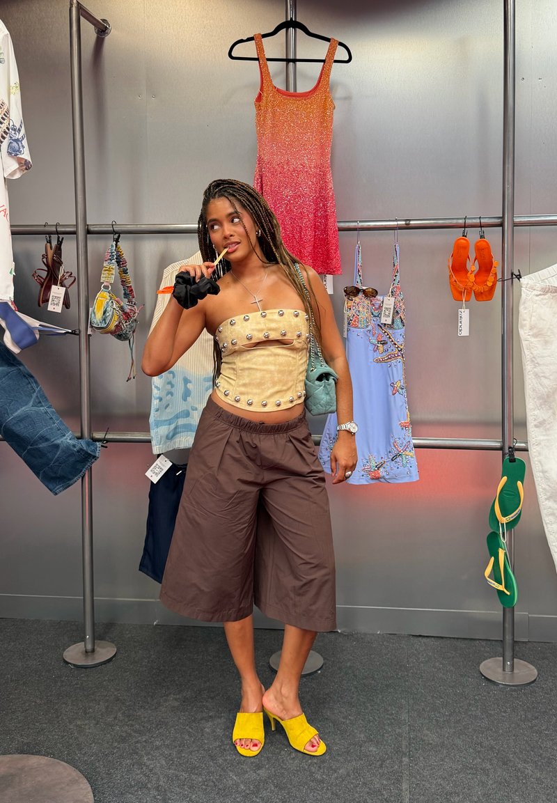 Femme posant dans un magasin portant un top tube beige clouté, un pantalon large marron, des talons jaunes, tenant un chouchou noir et un sac à main vert.