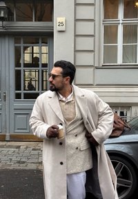 Lang beige kåpe over en strukturert kremfarget genser og hvite bukser. Brune hansker og holder en kaffekopp. Grå dør og bil i bakgrunnen.