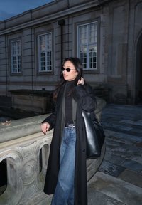 Black long coat, fitted black turtleneck, high-waisted blue jeans. Black tote bag, dark sunglasses. Standing by a stone railing in the evening light.