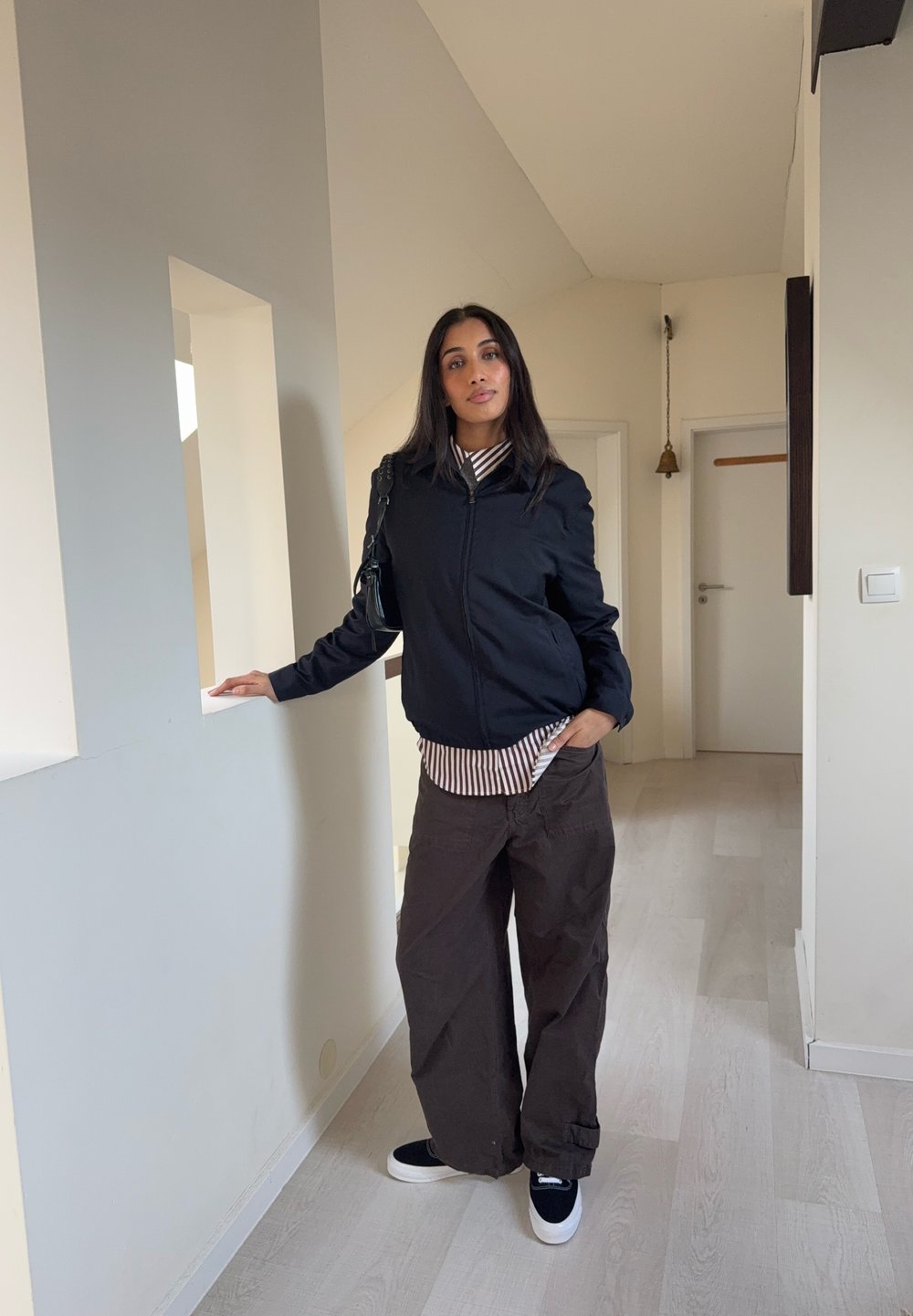 Veste bomber noire zippée portée sur un t-shirt rayé blanc et noir, associée à un pantalon marron ample et des baskets noires. Environnement intérieur avec des murs blancs.