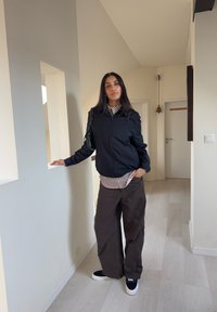 Veste bomber noire zippée portée sur un t-shirt rayé blanc et noir, associée à un pantalon marron ample et des baskets noires. Environnement intérieur avec des murs blancs.