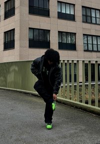 Oseba v črni jakni in svetlih zelenih čevljih se skloni, da prilagodi en čevelj na tlakovanem pločniku ob bež stavbi in ograji.