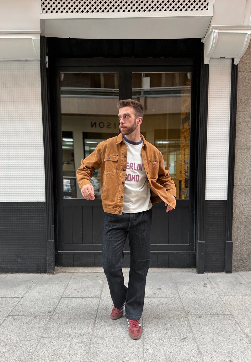 Homme portant une veste marron, une chemise blanche avec texte, un jean foncé et des chaussures rouges marchant sur un trottoir en ville devant des portes doubles noires.
