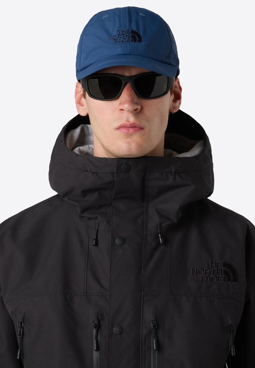 Blue zip-up jacket with a contrasting black pocket, complemented by matching blue trousers. It features a hood, a sporty design, and black trainers.
