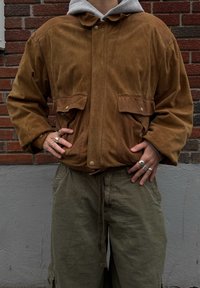 Osoba ubrana w brązową zamszową kurtkę na szarej bluzie z kapturem i oliwkowo-zielonych spodniach, stojąca z rękami na biodrach na tle ceglanej ściany.