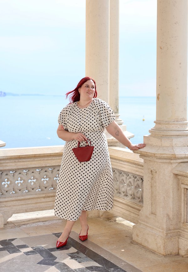 Frau mit roten Haaren in weißem Polka-Dot-Kleid hält eine kleine rote Handtasche und steht neben einer steinernen Balustrade mit Ozeanhintergrund.