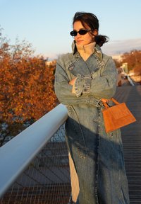 Jeans trenč plašč z ohlapnim krojem, svetlo modre barve in subtilnimi šivi, kombiniran z rjavim semiš torbico.