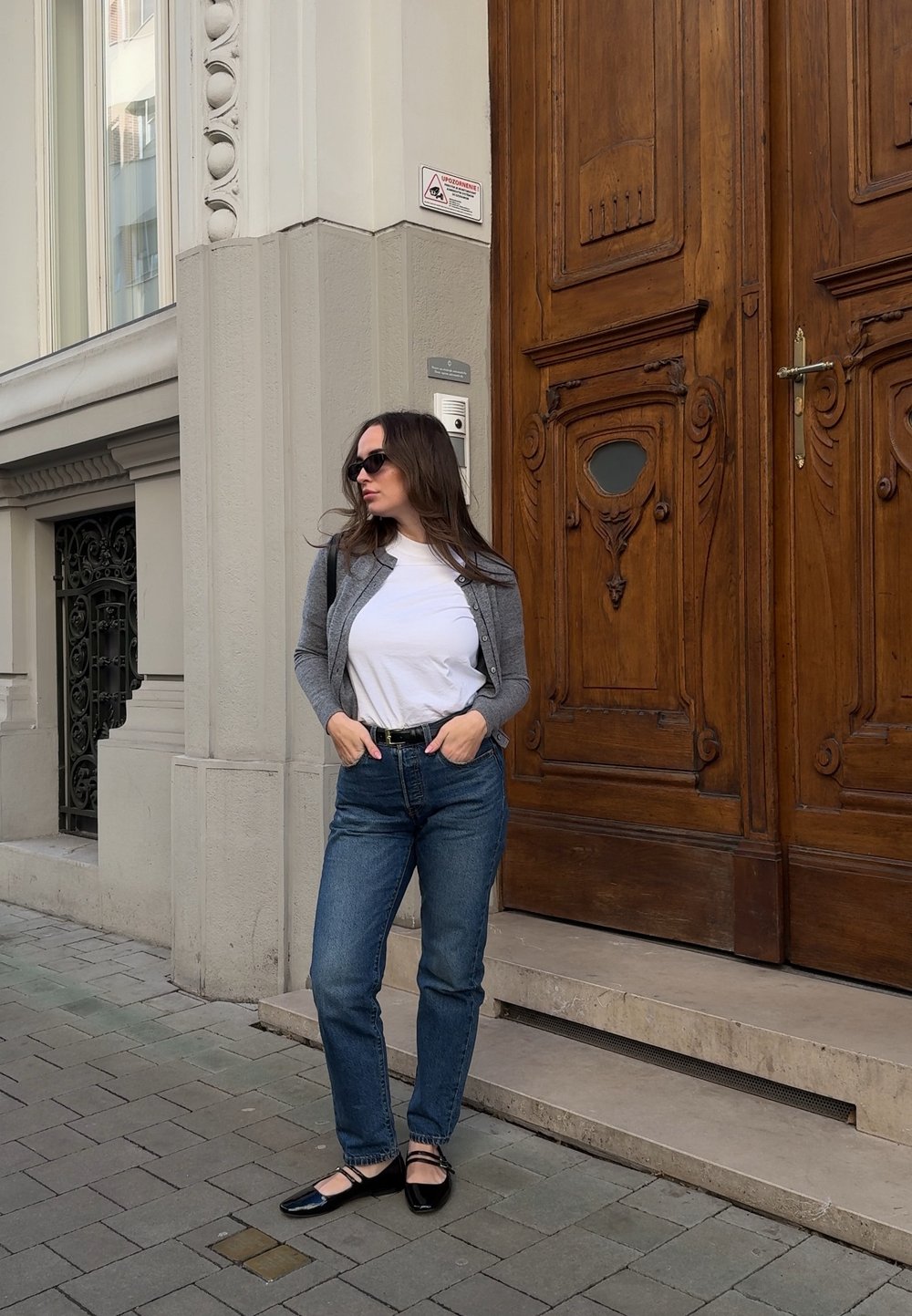 Γκρι πλεκτή ζακέτα πάνω από λευκό t-shirt, μπλε τζιν και μαύρες γόβες. Ξύλινη πόρτα με περίτεχνα σχέδια στο παρασκήνιο. Λιθόστρωτη πλακόστρωση.