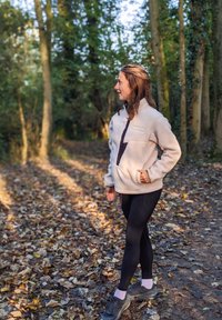 Pulôver de felpa em bege claro com meio zíper, bolso no peito e detalhes escuros, combinado com leggings pretas e ténis cinzentos num ambiente florestal.