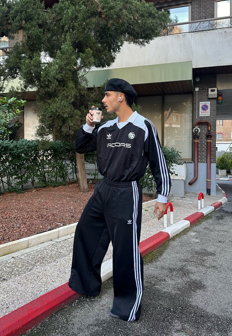 Μαύρο αθλητικό κοστούμι Adidas με λευκές ρίγες, διαθέτει κολάρο και λογότυπο, φορεμένο με μαύρο μπερέ. Το άτομο κρατάει ένα φλιτζάνι καφέ.