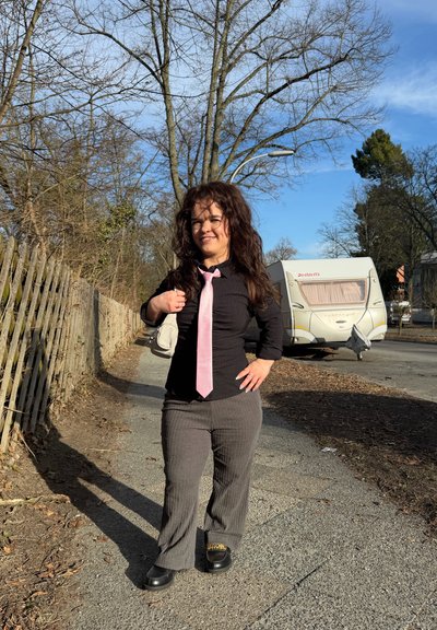 Mujer con cabello rizado que lleva una camisa negra, corbata rosa, pantalones grises y zapatos negros, caminando por un sendero junto a una valla de madera y una autocaravana estacionada.