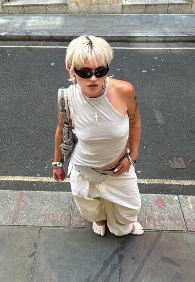 Persona rubia con top sin mangas blanco, pantalones anchos, zapatos de dedo, gafas de sol y accesorios plateados, de pie en la acera mirando hacia arriba.