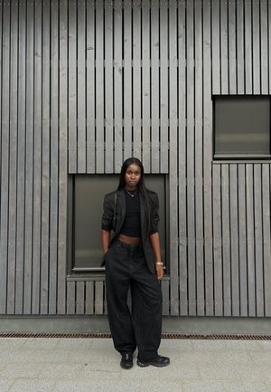 Svarta oversized denimbyxor och en åtsittande svart topp, lager på lager med en mörk blazer. Bakgrunden har vertikala träslipar.