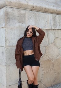 Giacca in suede marrone sopra un crop top a righe black e white, abbinata a pantaloni corti neri e stivali neri alti fino al ginocchio, su sfondo di una parete di pietra.