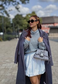 Palton oversized gri, pulover tricotat gros, fustă albă strălucitoare și geantă gri cu detalii din dantelă. Ochelarii de soare negri completează aspectul.