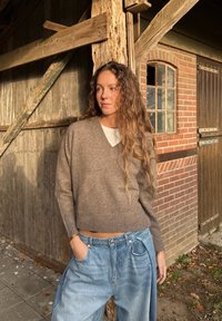 Brązowy sweter z dzianiny z dekoltem w serek i prążkowanym obszyciem, zestawiony z jasnoniebieskimi, oversize spodniami. Rustykalne drewniane tło z ceglaną ścianą.