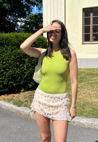 Zöld egyrészes fürdőruha aszimmetrikus dekoltázzsal, világos rózsaszín, többrétegű, fodros mini szoknyával párosítva. Átlátszó kézitáska is jár hozzá.