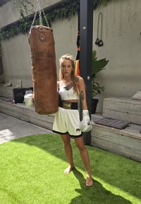 Μπλέντζα από καφέ δέρμα κρέμεται από αλυσίδα; άτομο με λευκή αμάνικη μπλούζα και μαύρες σορτς φορά λευκά γάντια πυγμαχίας, στέ stands σε πράσινο χόρτο.