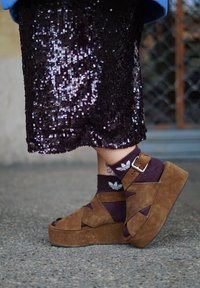 Alakehad pruunide seemisnahast platvormsandaalidega, mis on kantud lilla Adidas sokkide peale, koos musta litritega seelikuga ja nähtava pahkluu tätoveeringuga.