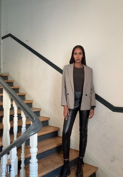 Blazer gris a cuadros sobre una parte superior oscura, combinado con pantalones de cuero negros. La modelo está de pie sobre unas escaleras de madera con barandas blancas y un pasamanos oscuro.