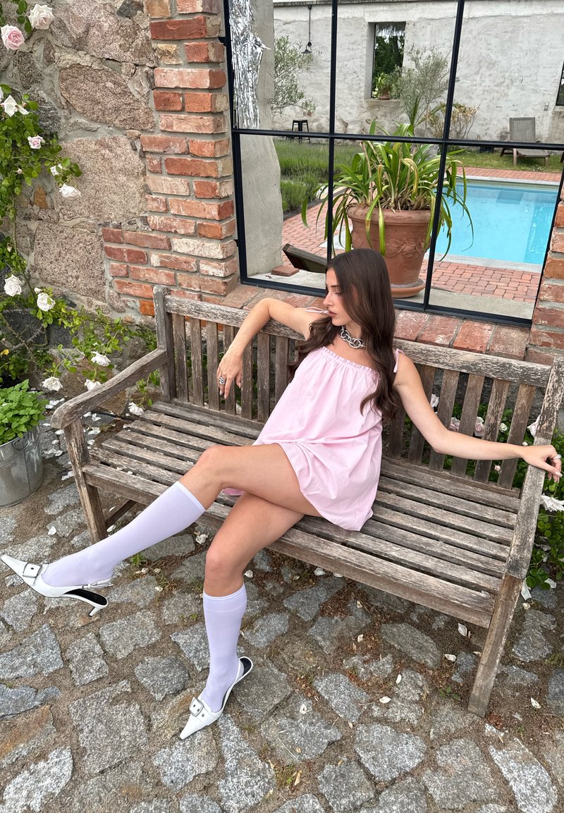 Femme en robe rose et chaussettes blanches montantes jusqu'aux genoux assise sur un banc en bois près d'un mur en briques avec une fenêtre montrant une plante en pot et une piscine à l'extérieur.