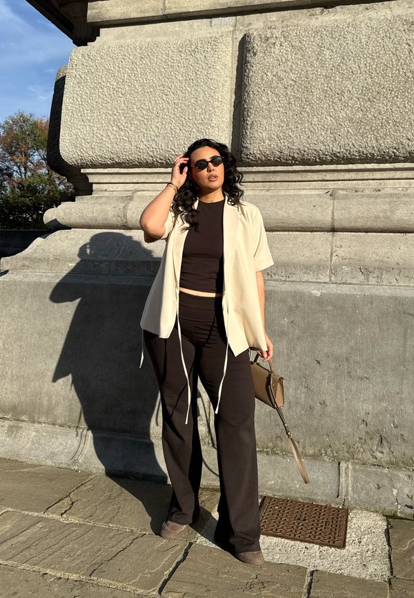 Light cream short-sleeve shirt over a black crop top and black wide-leg trousers. Holding a small brown handbag; wearing dark sunglasses.