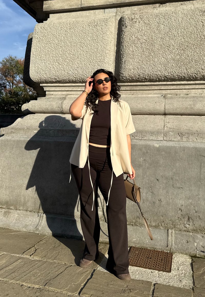 Camisa de manga curta de creme clara sobre um top cropped preto e calças largas pretas. A segurar uma pequena bolsa castanha; a usar óculos de sol escuros.