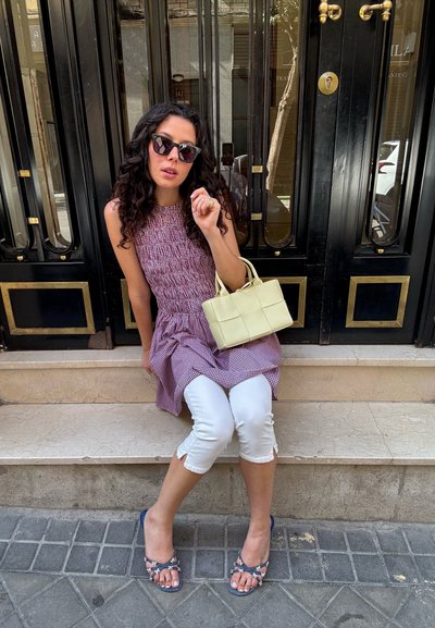 Vestido de tirantes con smock y patrón de cuadros morados, combinado con pantalones cortos blancos y sandalias florales. Sosteniendo un bolso estructurado de color amarillo pálido.