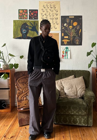 Chaqueta negra corta, pantalones de pierna ancha a rayas grises y zapatos negros. La habitación cuenta con muebles de madera, plantas verdes y varias obras de arte en las paredes.