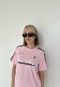 Maglietta rosa con logo "adidas" nero e grafica a stella, con strisce nere lungo le maniche. Indossata con occhiali da sole neri.