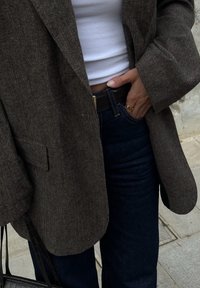 Brun fiskbensmönstrad blazer över en vit ribbad topp, kombinerad med mörka denimjeans. En svart handväska hålls i vänster hand.