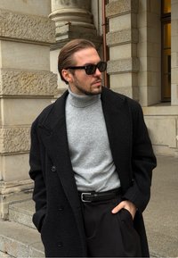 Manteau en laine noire porté sur un pull à col roulé gris, associé à un pantalon noir. La personne porte des lunettes de soleil foncées et a les cheveux coiffés en arrière.