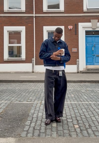 Chaqueta de denim con cuello, pantalones negros oversized y zapatos marrones; sosteniendo una bebida, de pie sobre un pavimento de adoquines cerca de una puerta azul.