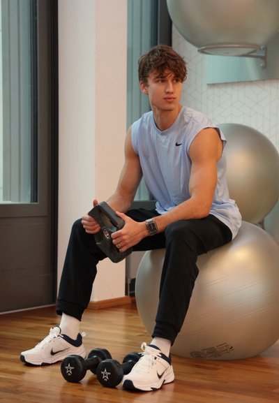 Camiseta sin mangas azul claro, pantalones negros, zapatillas blancas con detalles en azul marino, sosteniendo una mancuerna negra, sentado sobre una pelota de ejercicio plateada.