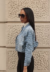 Nő, hosszú sötét hajjal, oversized fekete napszemüveget visel, mintás kék farmerdzseki, fehér garbó és fekete nadrág van rajta, oldalra áll.