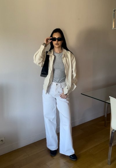 Mujer vestida con una chaqueta beige, camiseta gris, pantalones anchos blancos, zapatos negros, gafas de sol y llevando un bolso negro de hombro, de pie en el interior junto a una mesa de cristal.